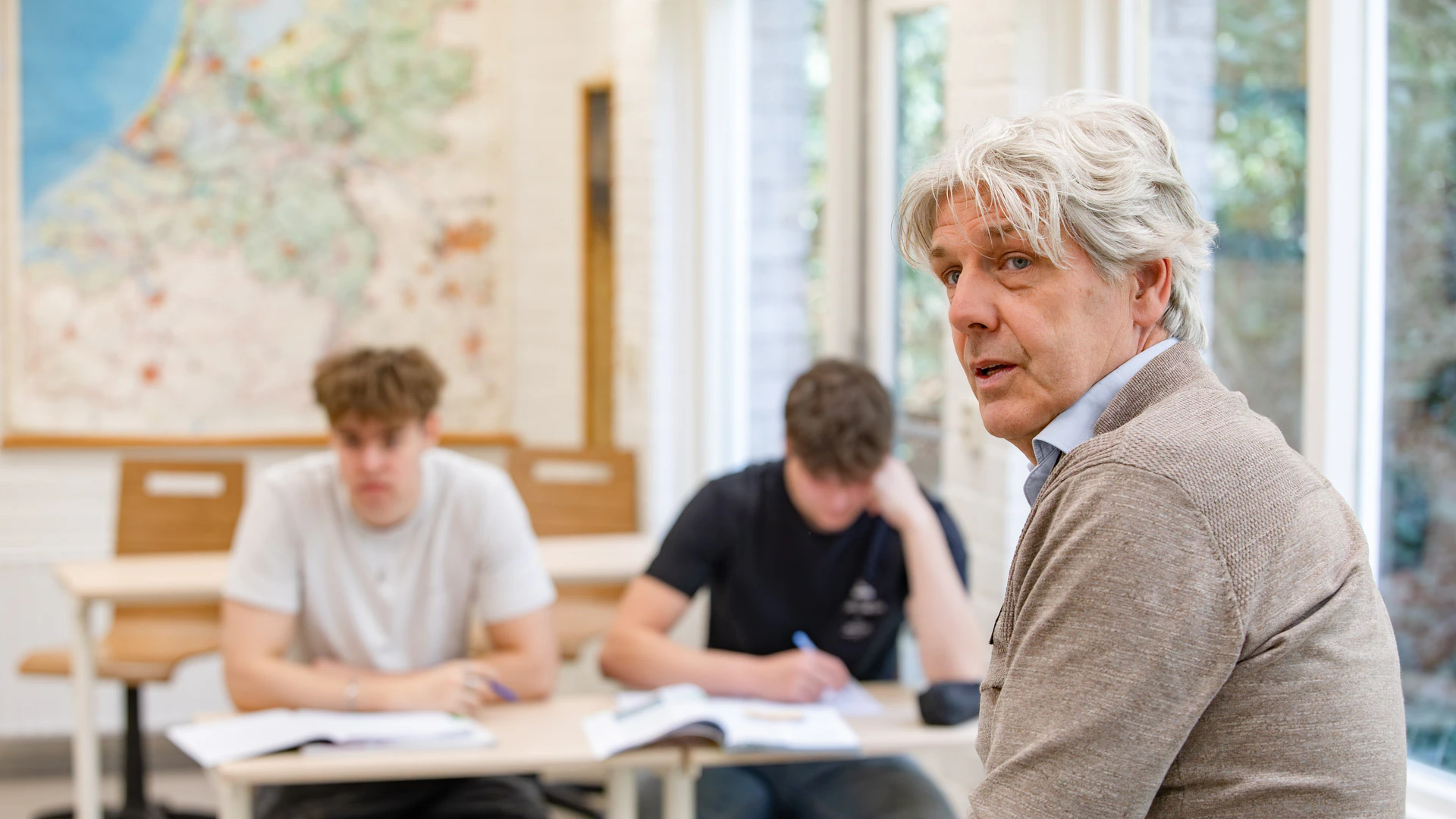 Een meneer in een klas lokaal kijkt weg van de camera. Op de achtergrond 2 schrijvende jongens.