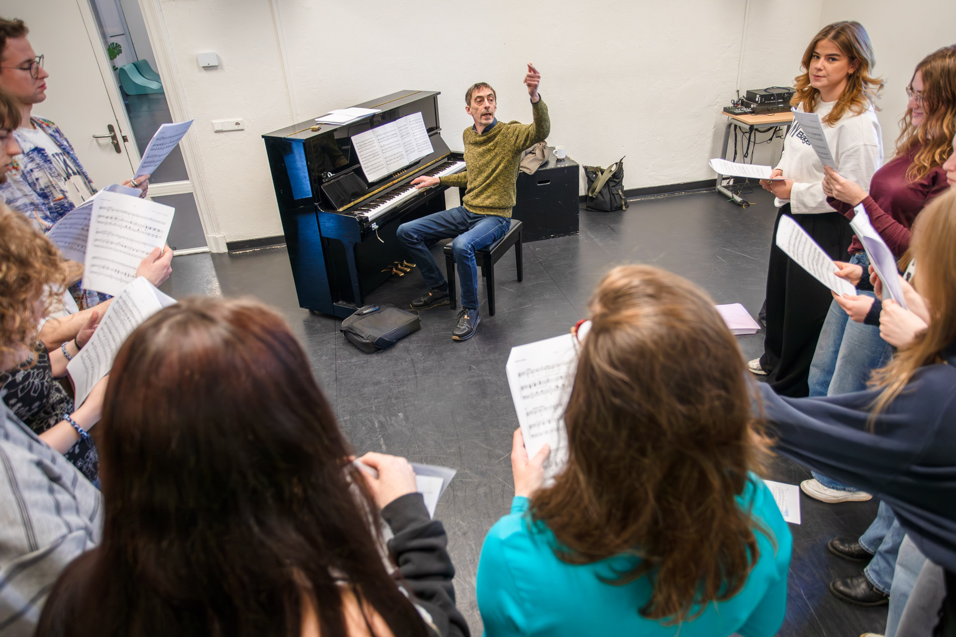 Een man zit achter een piano en geeft instructies aan zijn studenten.