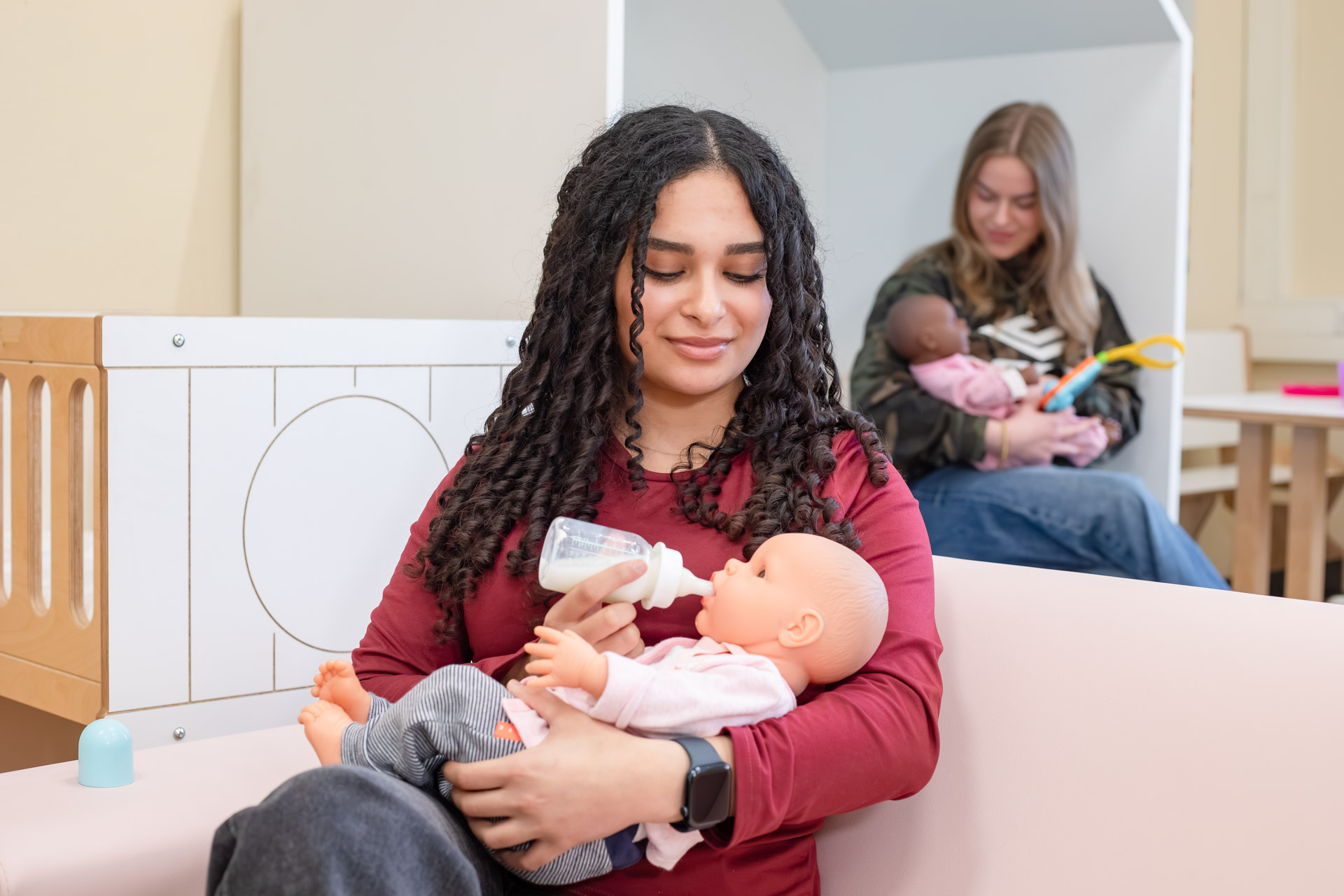 Meisje heeft een babypop in de hand die ze een flesje drinken geeft. Op de achtergrond zie je een ander meisje met een babypop in de hand