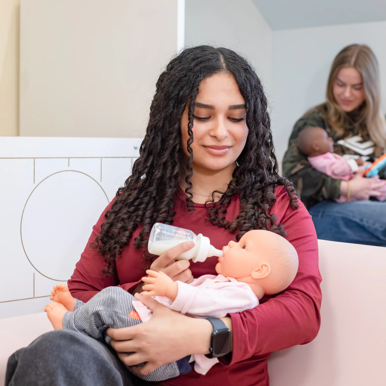 Meisje heeft een babypop in de hand die ze een flesje drinken geeft. Op de achtergrond zie je een ander meisje met een babypop in de hand