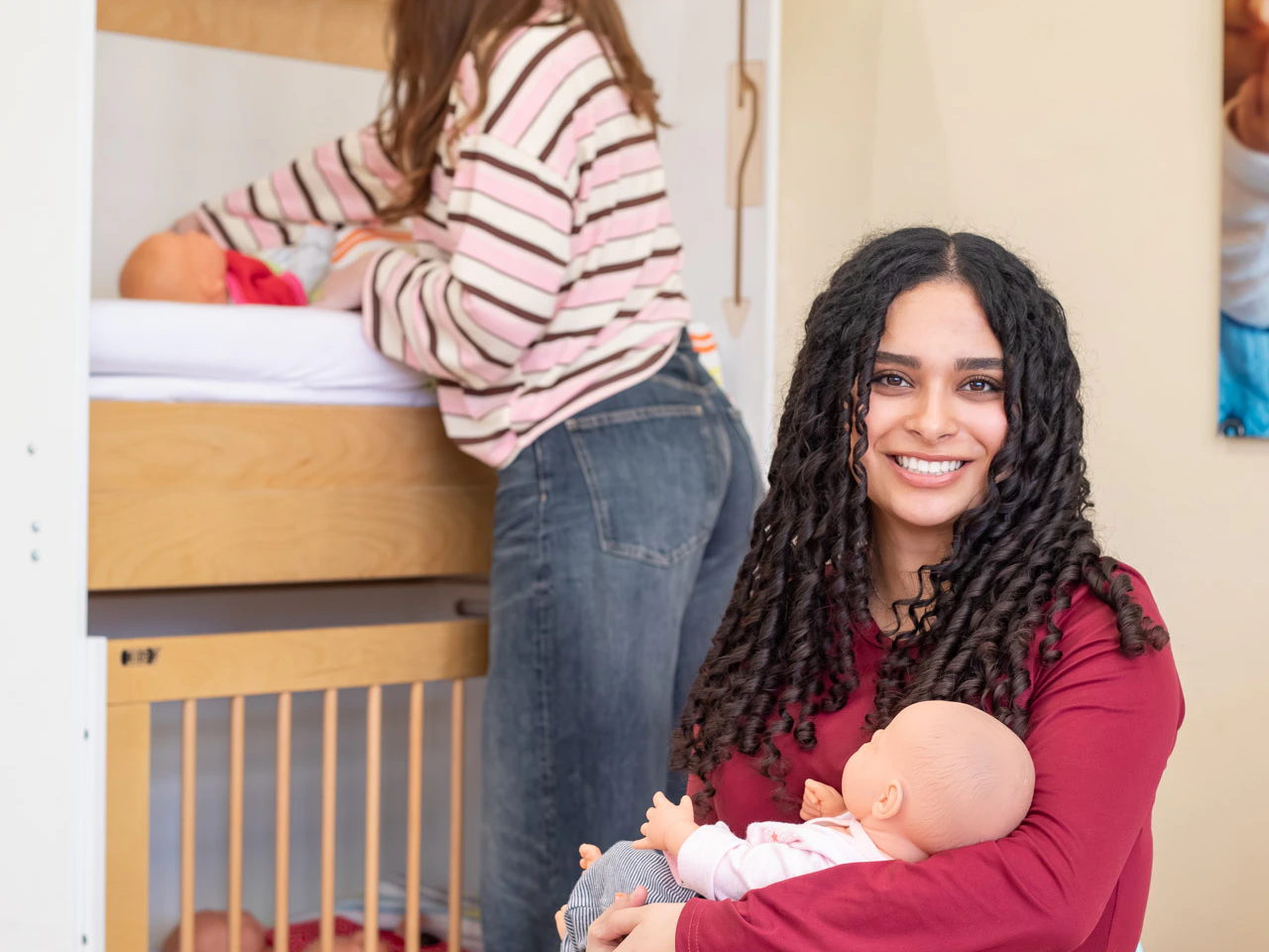 Meisje heeft een babypop in de hand terwijl op de achtergrond een andere meisje een babypop aan het verschonen is