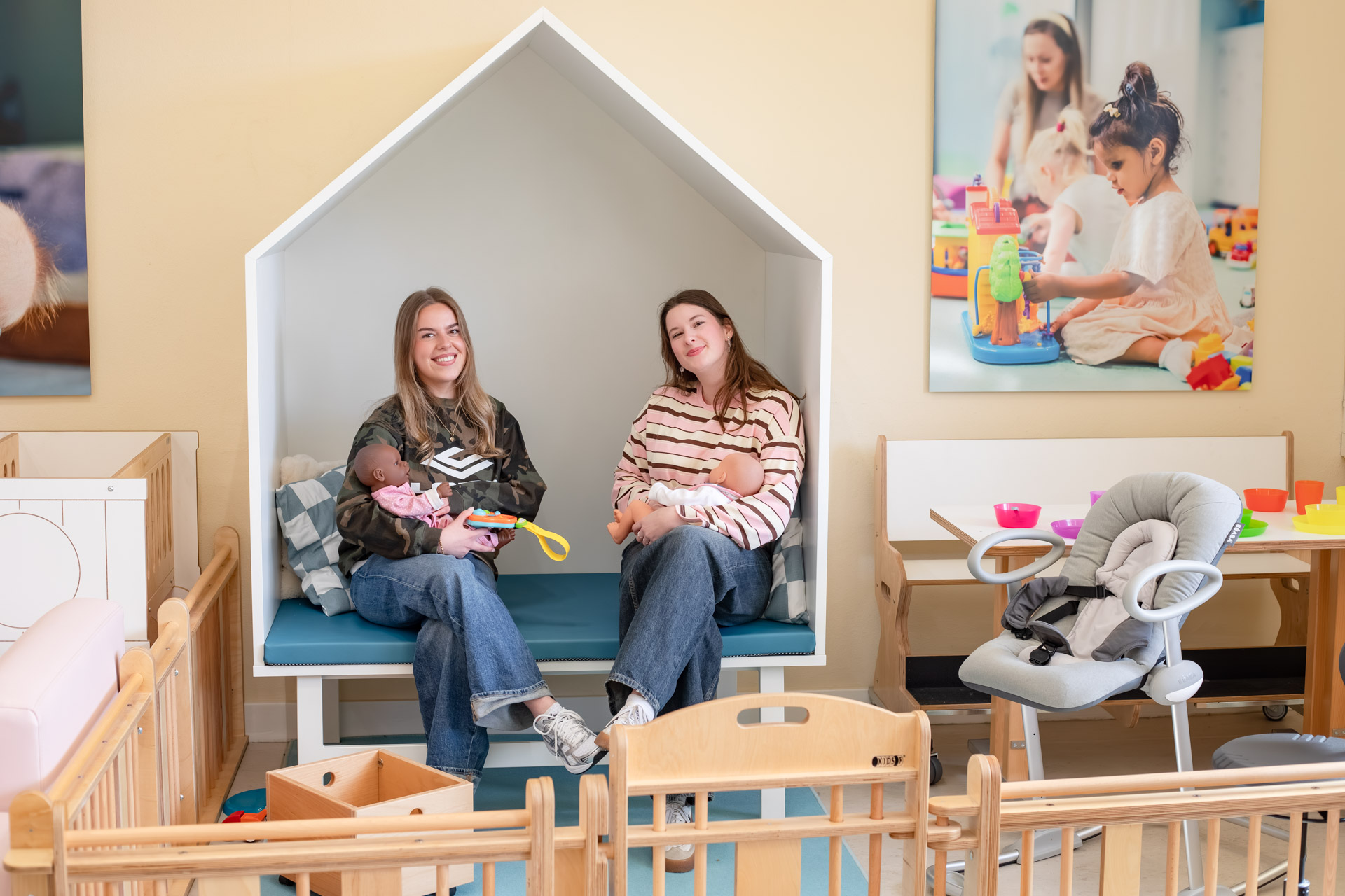 Je ziet 2 meisjes op een bank met allebei een babypop in de hand en kijken in de camera