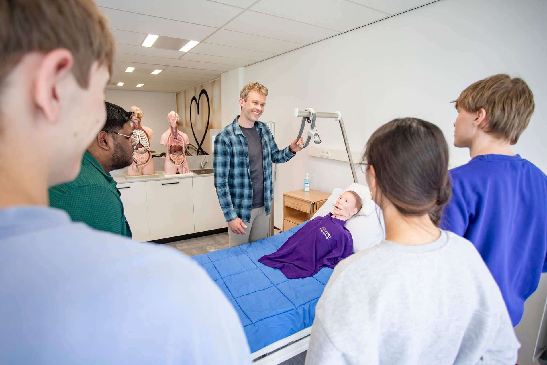 Man staat naast een ziekenhuisbed waar een pop ligt met aan de andere kant van het bed studenten