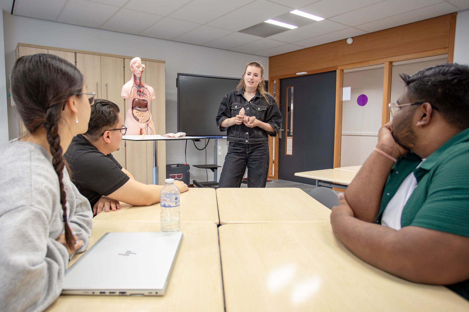 Vrouw staat voor een groep studenten en geeft les. Naast haar zie je anatomisch torsomodel staan