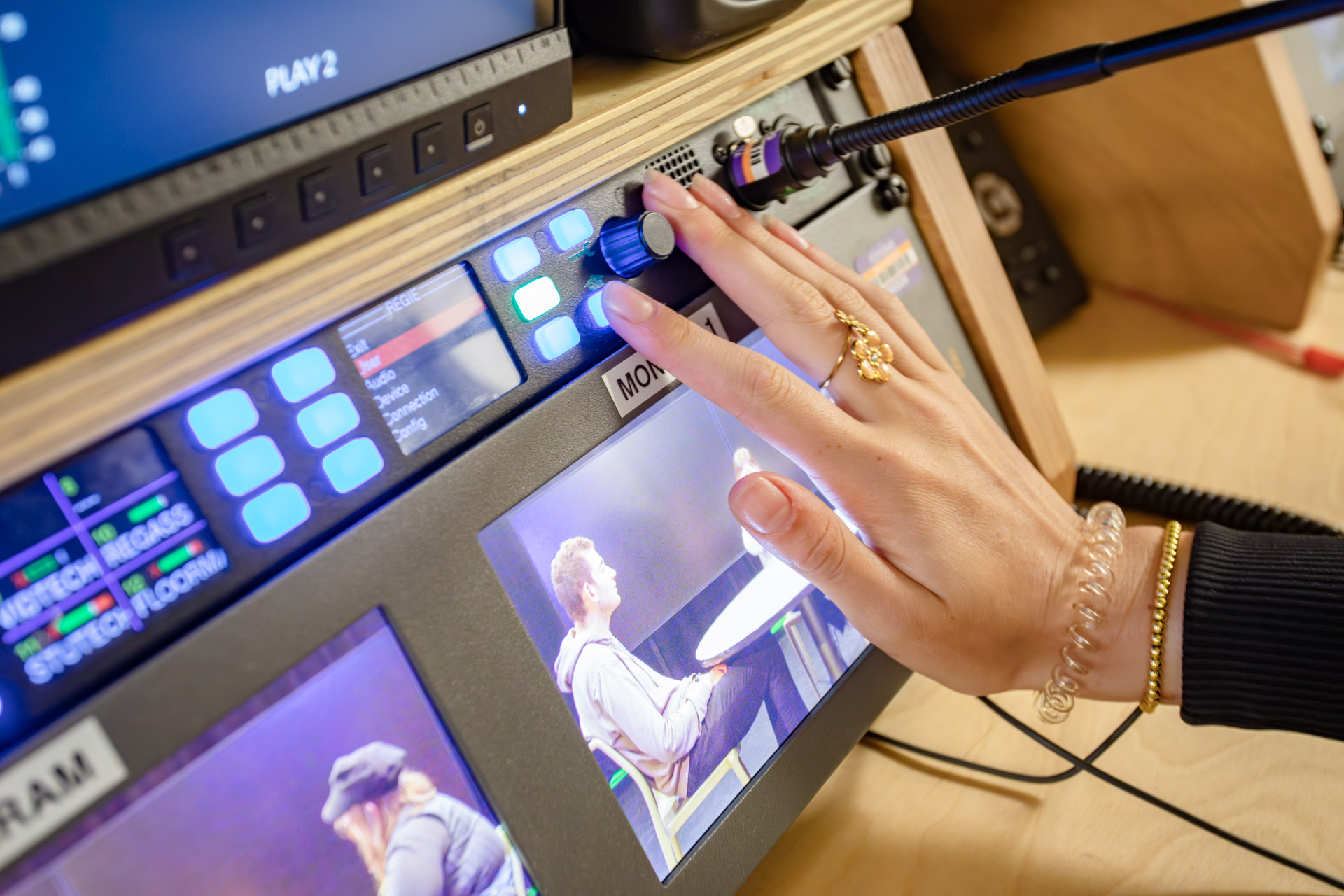 Je ziet een hand die aan een knop draait op het scherm van een televisiestudio. Op dat scherm zie je de camerabeelden