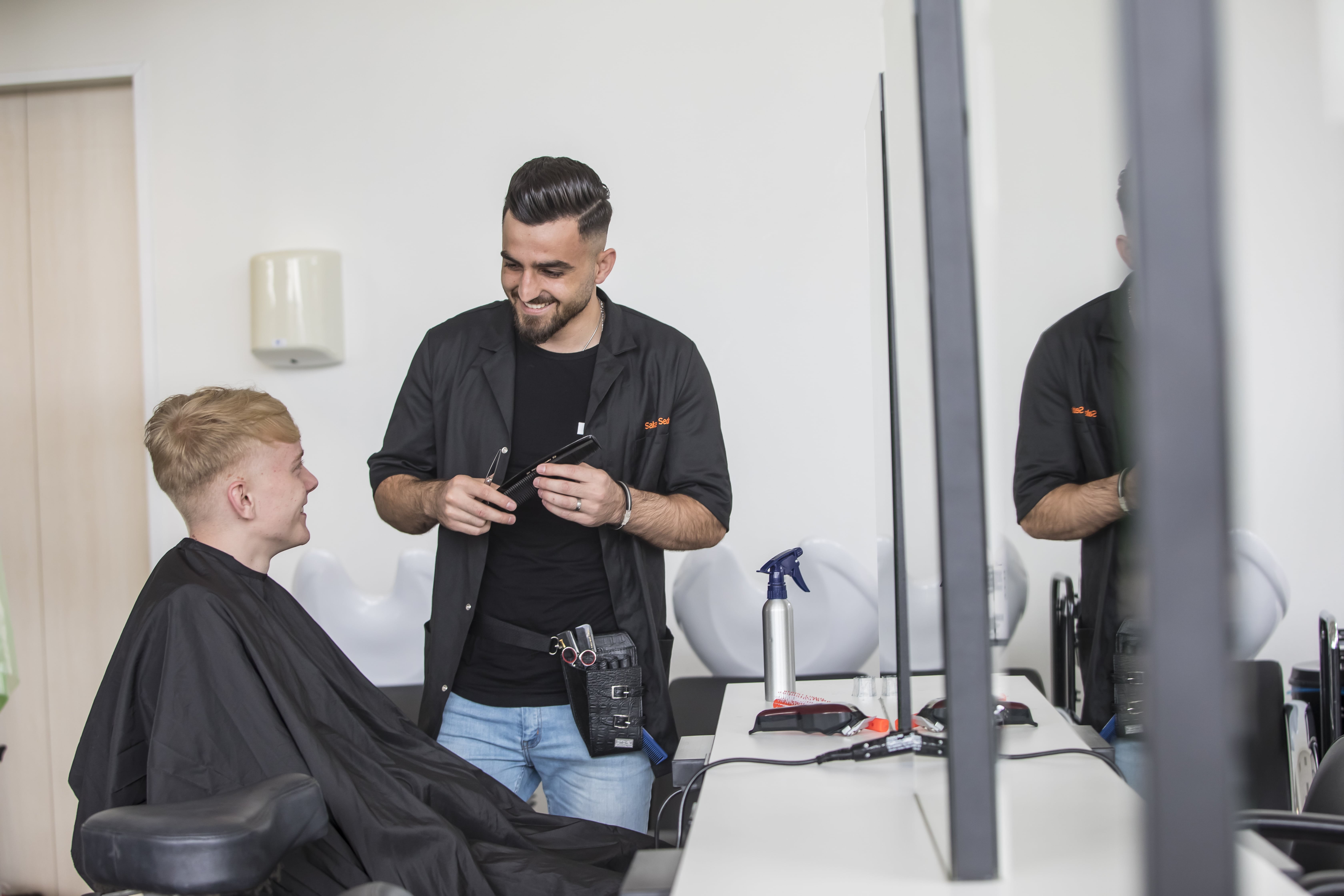 Student zit op de kappersstoel terwijl er een student naast staat