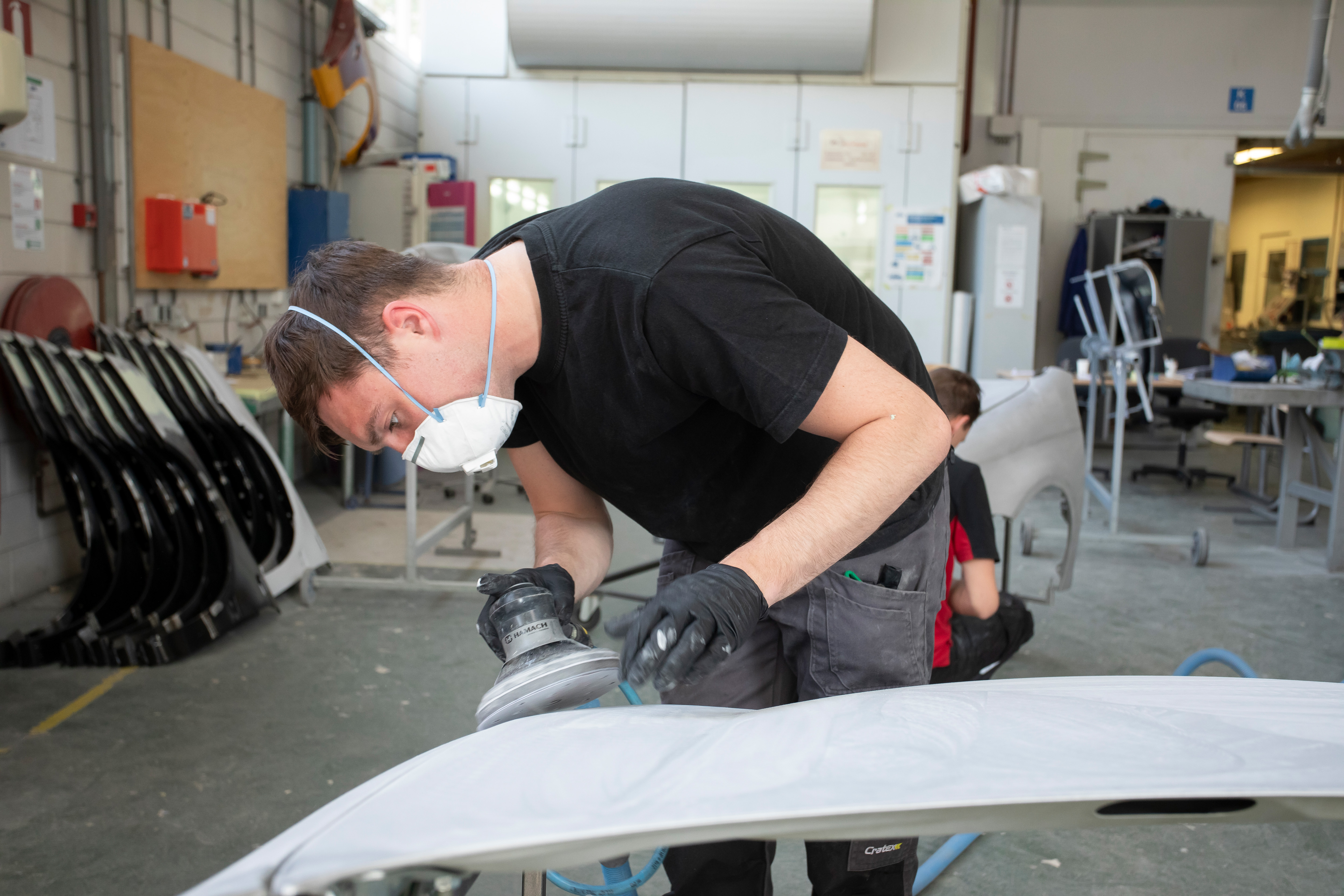 Jongen met een mondkapje op en werkkleding aan is een onderdeel van de auto aan het schuren met een scchuurapparaat