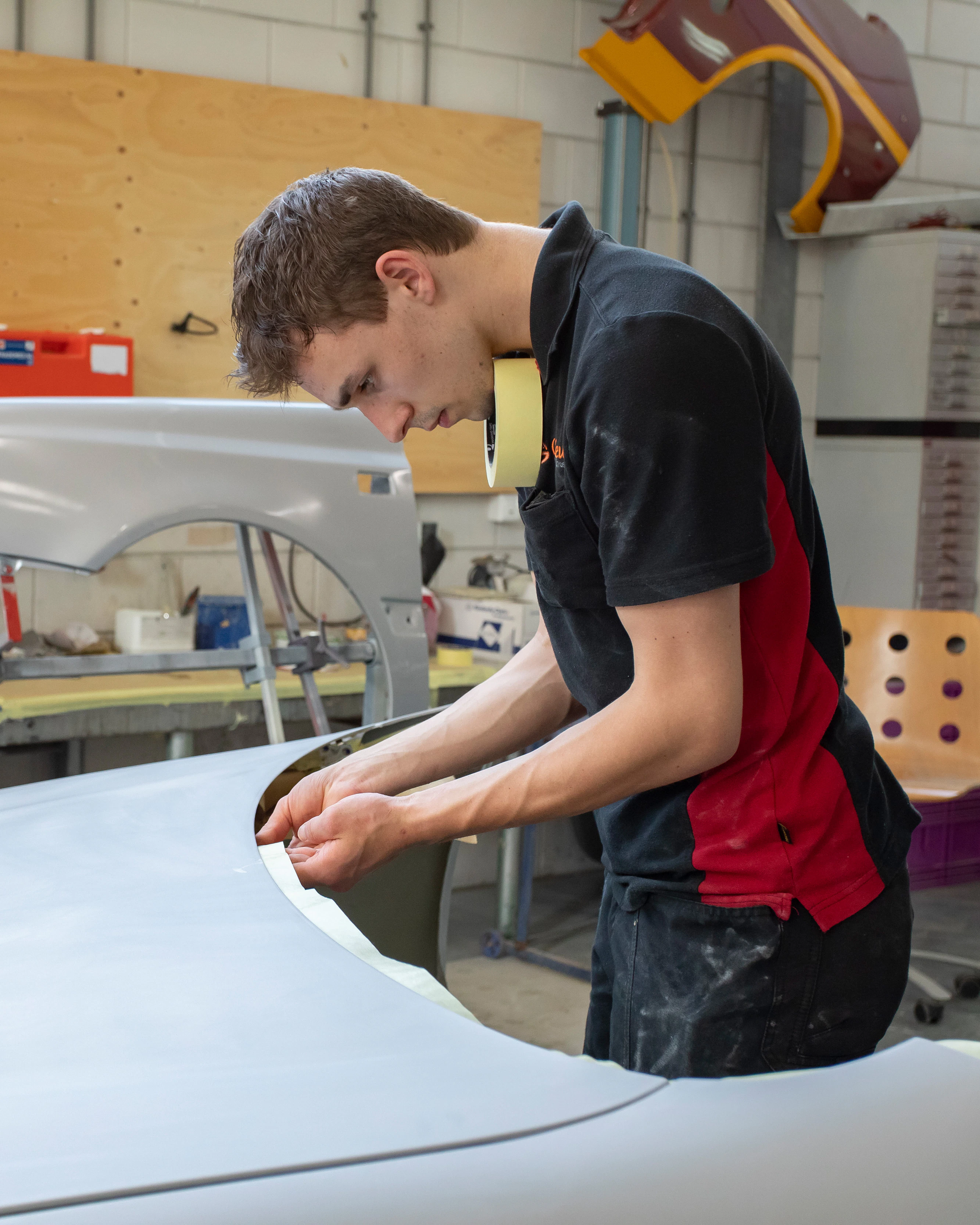 Jongen in werkkleding plakt tape op een onderdeel van de auto terwijl hij in de werkplaats staat