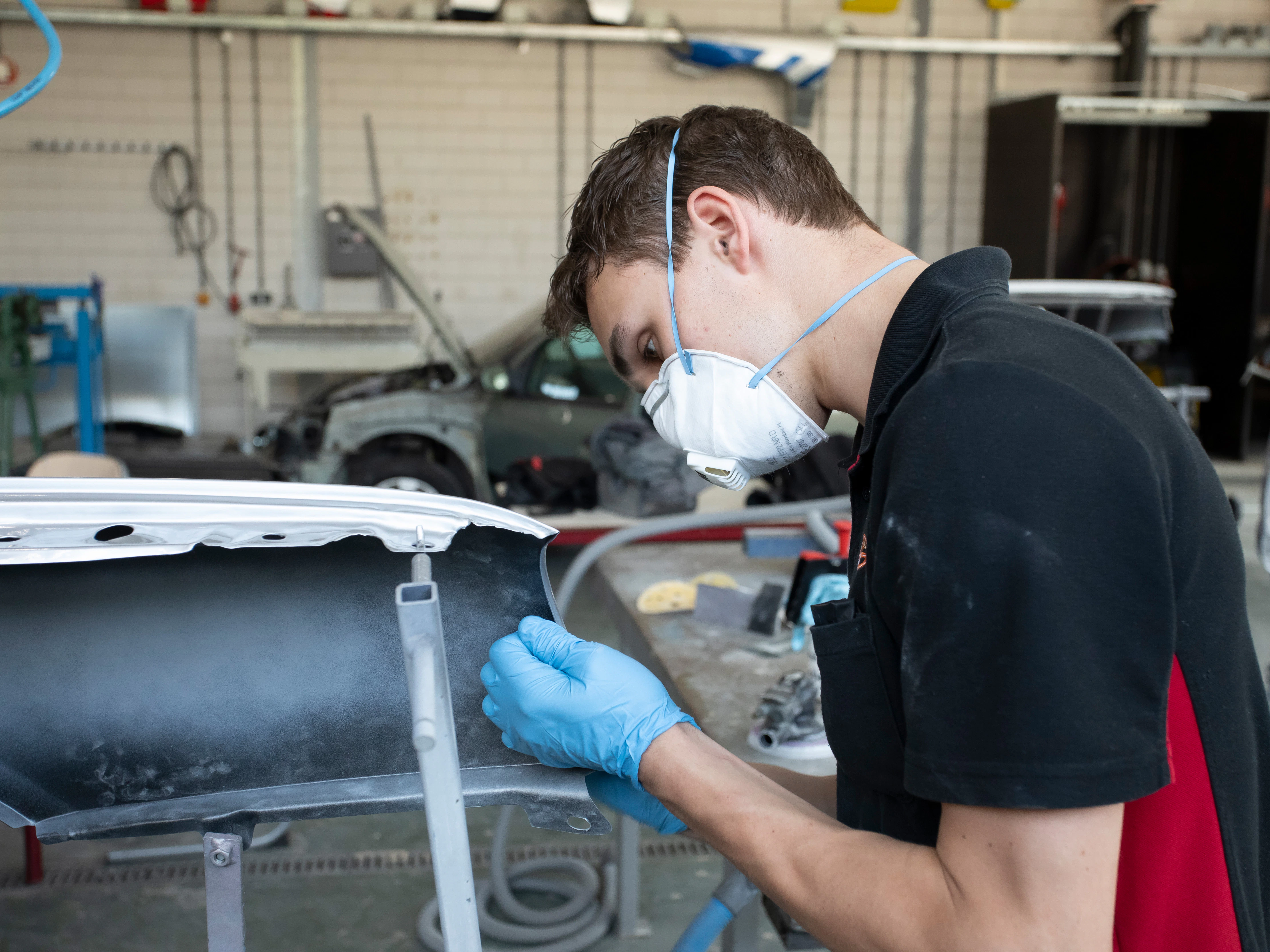 Jongen met een mondkapje op staat in de werkplaats en is bezig met een onderdeel van de auto