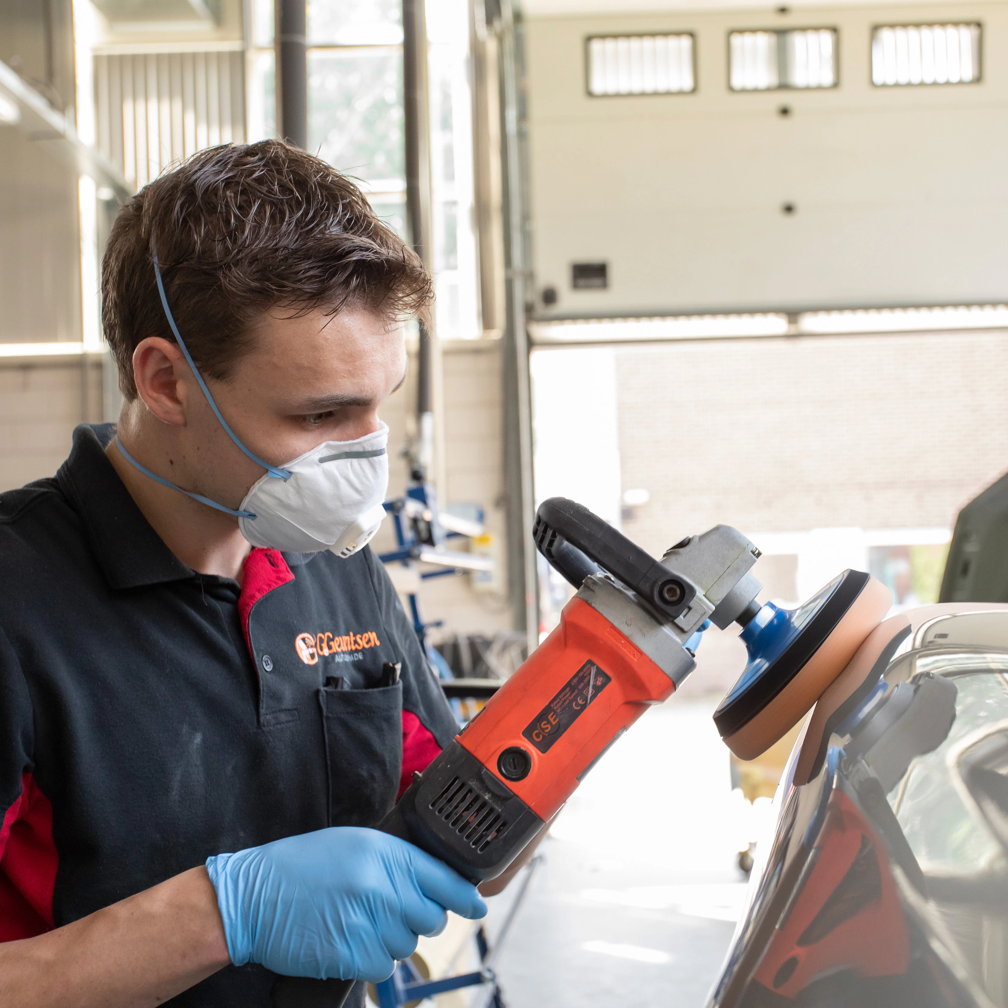 Jongen met een mondkapje op heeft een apparaat in de hand waarmee hij een onderdeel van de auto aan het boenen is