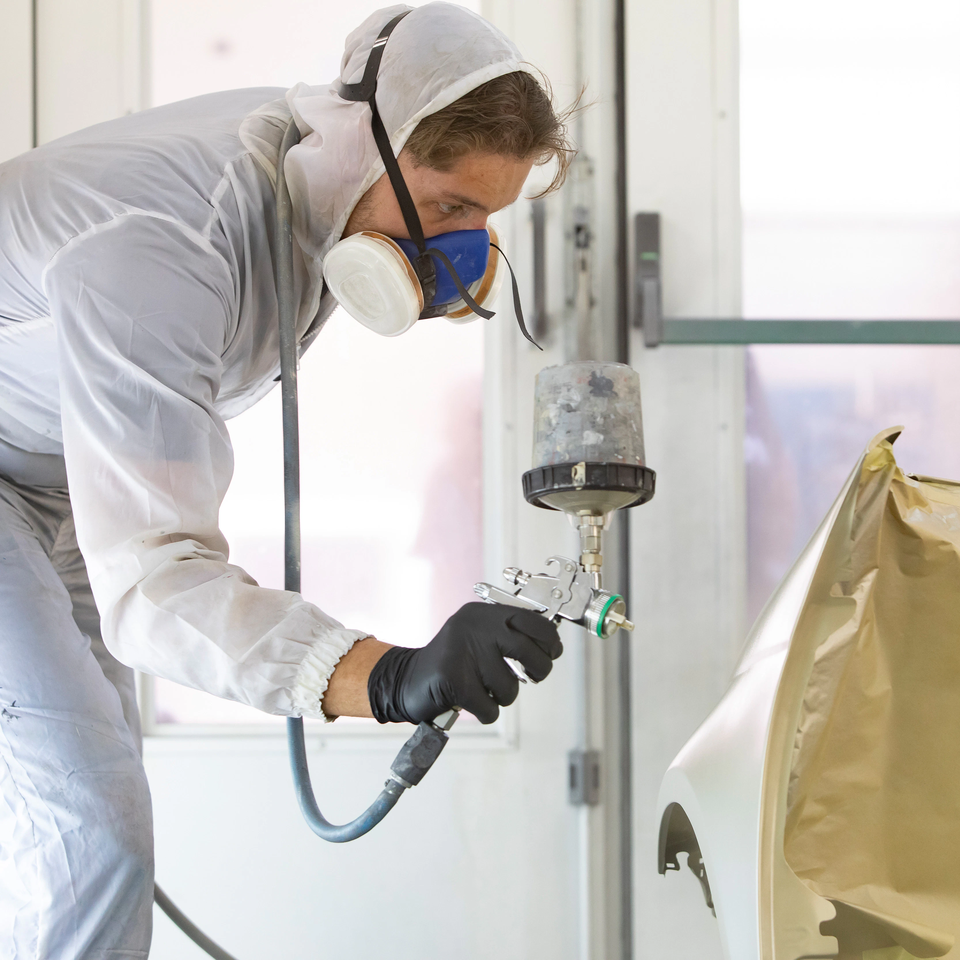 Jnogen in een wit pak heeft een spuitapparaat in de hand waarmee hij een onderdeel van de auto aan het spuiten is
