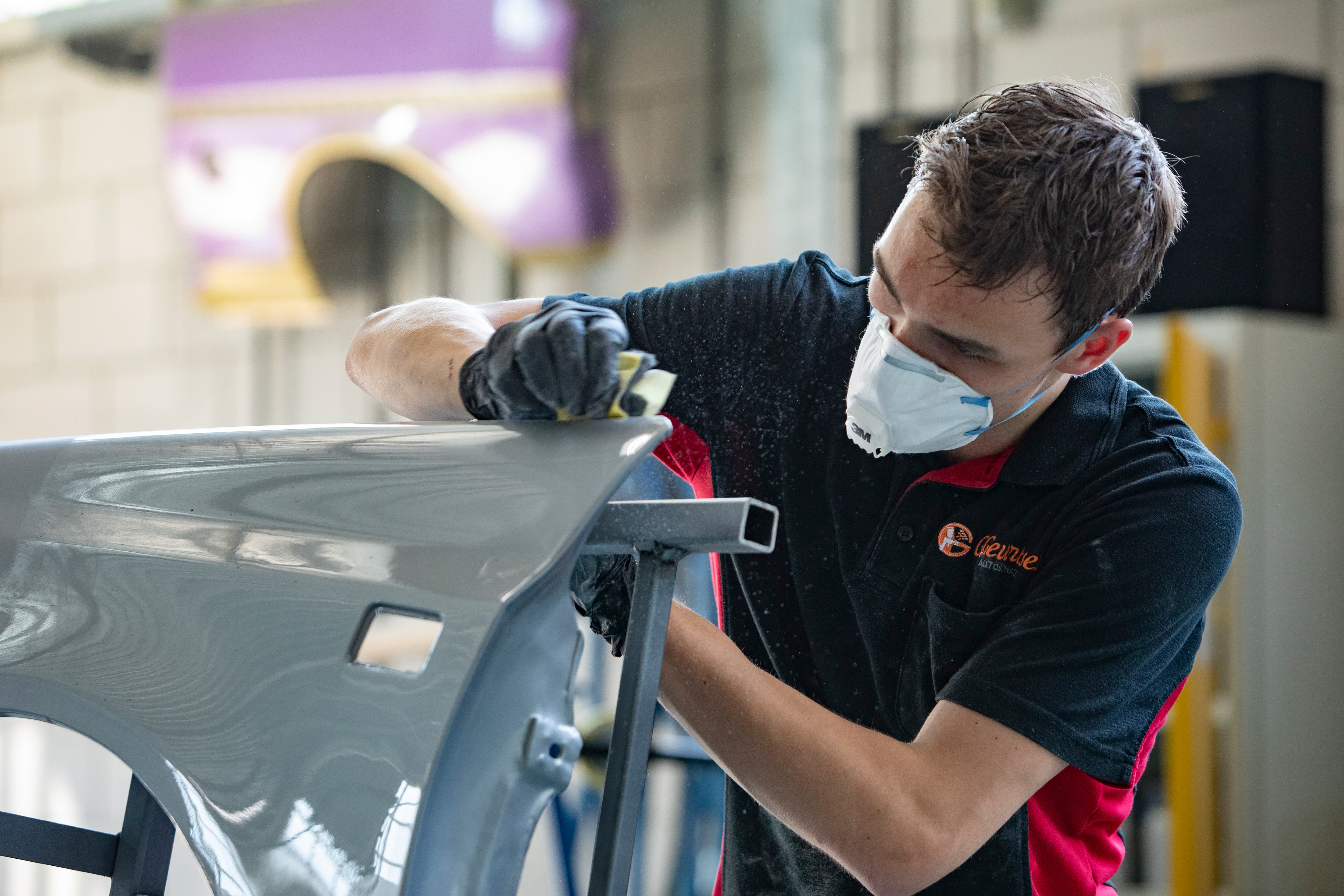 Jongen met een mondkapje op staat in de werkplaats. Hij veegt met een doekje over een onderdeel van de auto