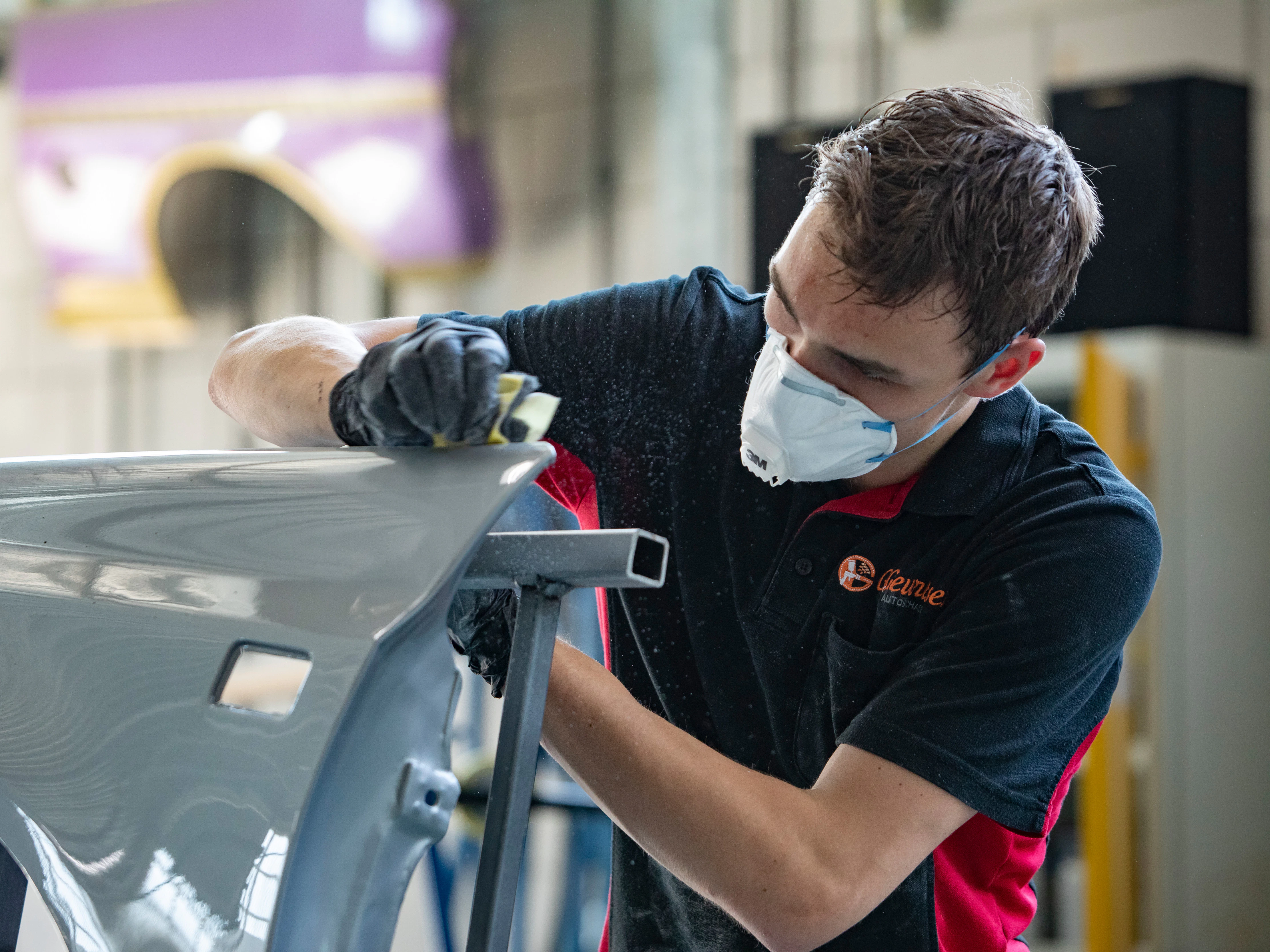 Jongen met een mondkapje op staat in de werkplaats. Hij veegt met een doekje over een onderdeel van de auto