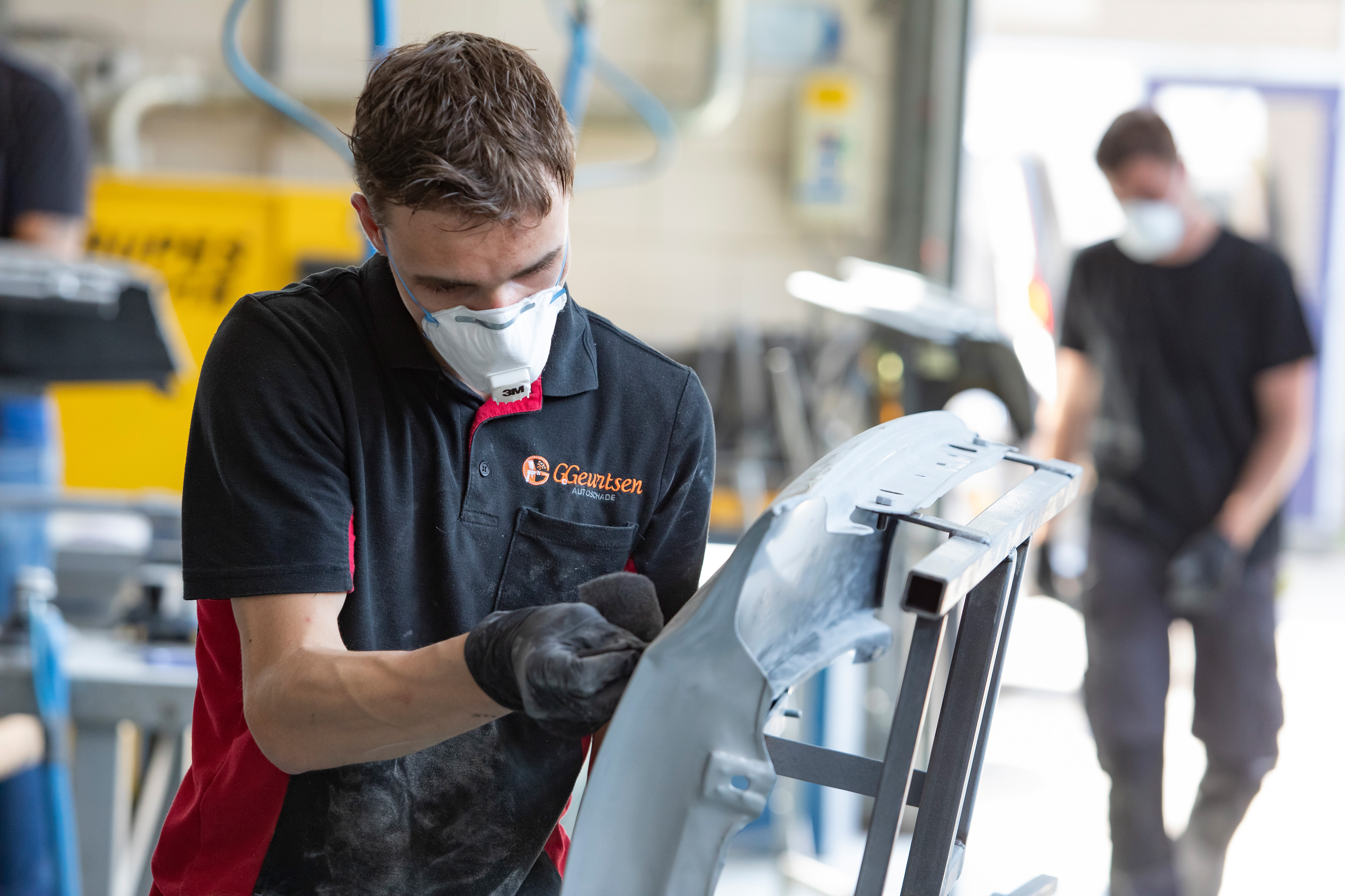 Jongen met een mondkapje op en werkkleding aan is een onderdeel van de auto aan het schuren met boenen