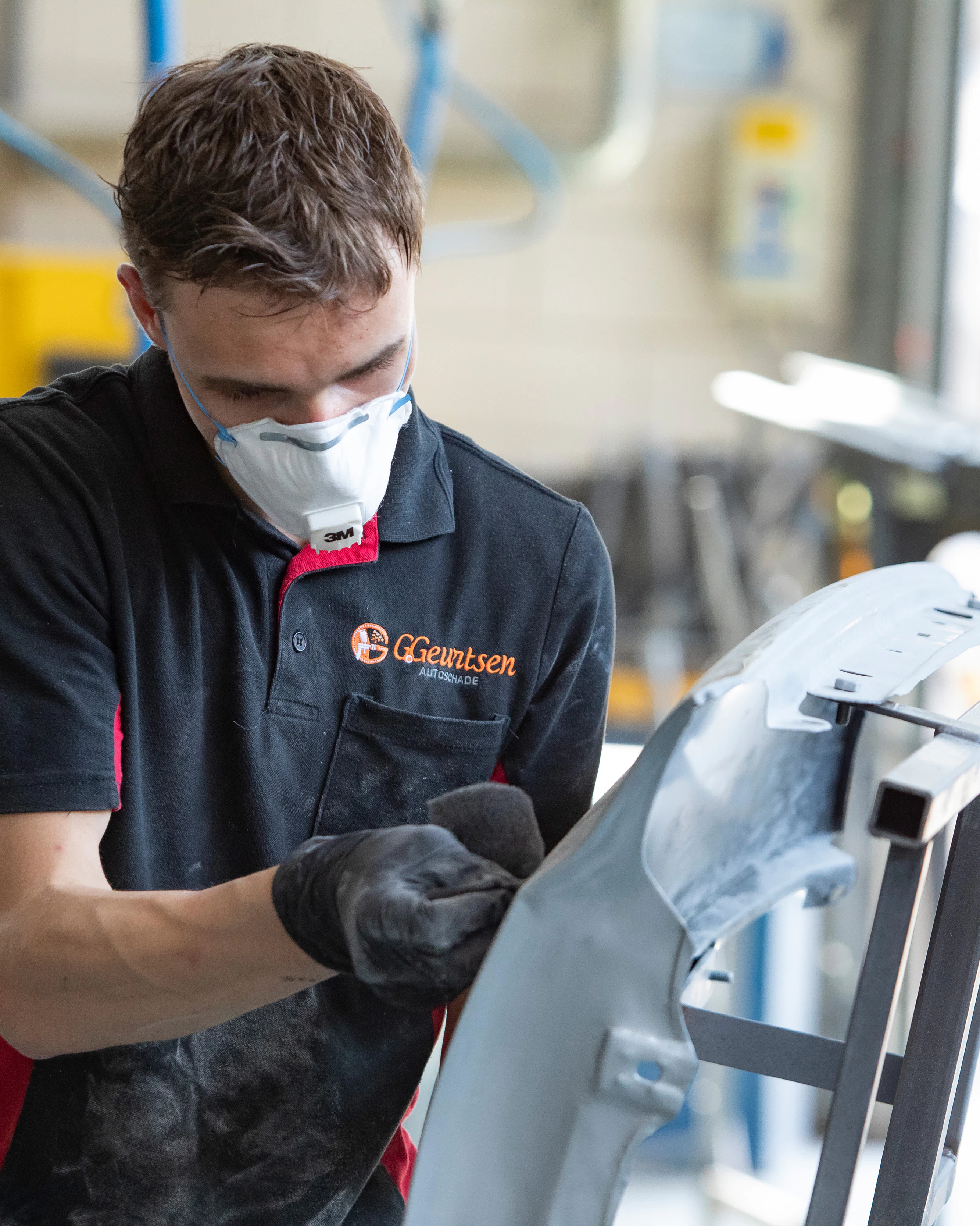 Jongen met een mondkapje op en werkkleding aan is een onderdeel van de auto aan het schuren met schuurpapier