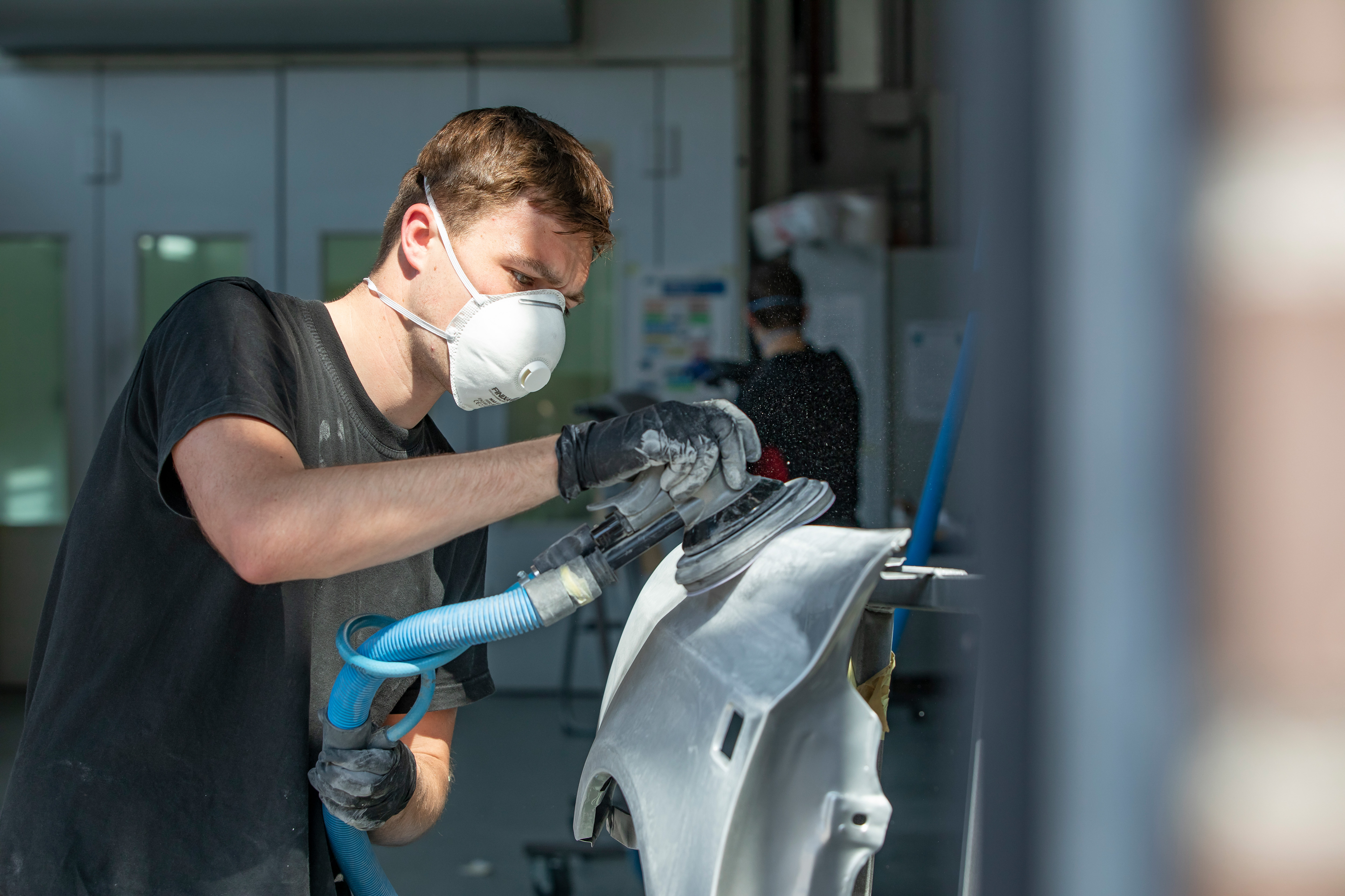 Jongen met een mondkapje op heeft een apparaat in de hand waarmee hij een onderdeel van de auto aan het schuren is