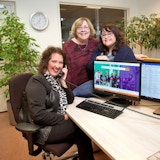 3 personen kijken in de camera terwijl ze achter een computer zitten. Een vrouw heeft een telefoon in de hand