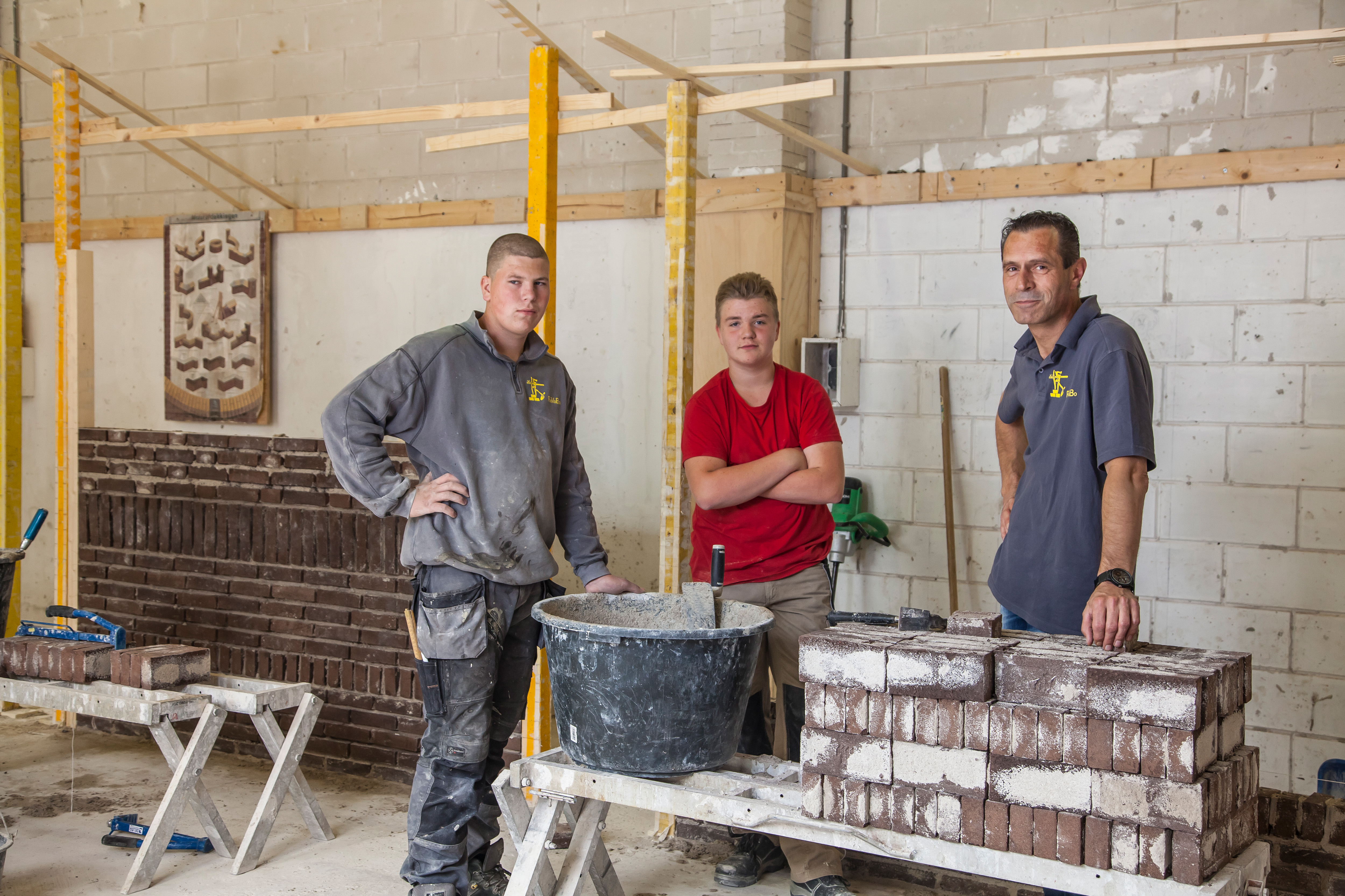 Twee studenten en docent staan bij gemetseld muurtje en kijken naar camera